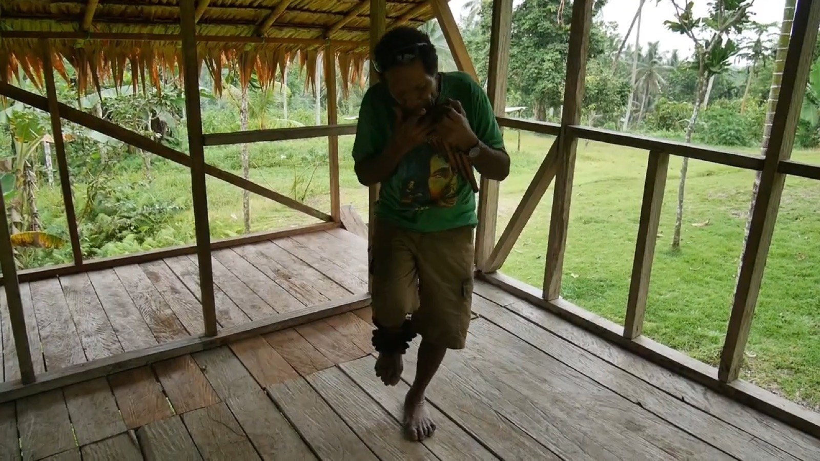 An old man with rattles around his ankle lifts one foot while playing a panpipe